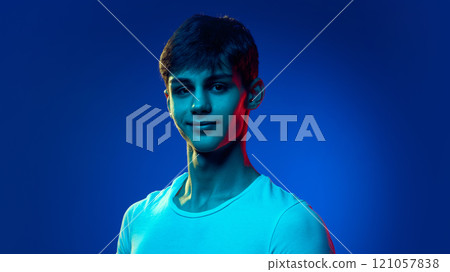 Portrait of handsome young man in white t-shirt posing with smile against blue studio background in neon light 121057838