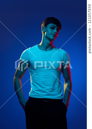 Portrait of handsome young man in white t-shirt, with muscular, sportive body, posing with smile against blue studio background in neon light Portrait of handsome young man in white t-shirt, with muscular, sportive body, posing with smile against blue studio background in neon light 121057839