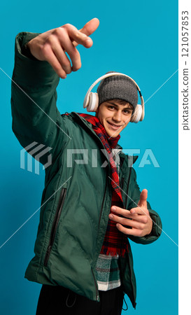 Young smiling man wearing winter puff jacket, red checkered scarf and gray hat, listening to music in headphones against blue studio background 121057853