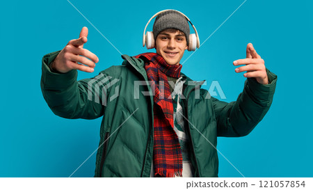 Young handsome man wearing winter puff jacket, red checkered scarf and gray hat, listening to music in headphones against blue studio background 121057854