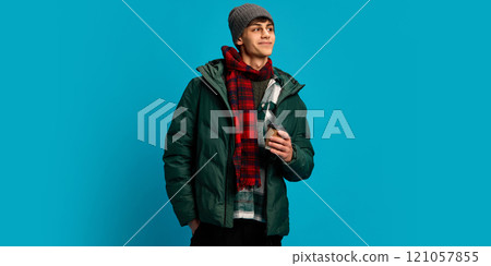 Young handsome man wearing winter puff jacket, red checkered scarf and gray hat, smiling,man holding cup of coffee against blue studio background 121057855