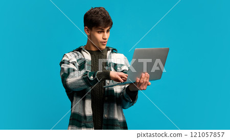 Emotional young handsome man, student in casual clothes holding laptop and looking with excited face against blue studio background 121057857