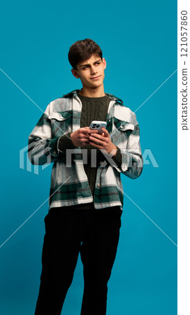 Portrait of young handsome man in casual clothes holding smartphone and texting message with contemplation against blue studio background 121057860
