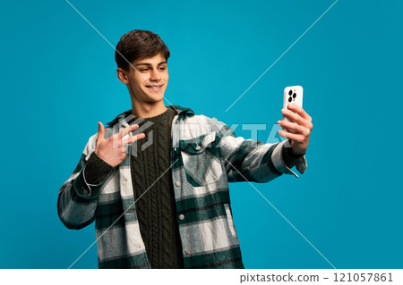 Handsome young guy in casual clothes, checkered shirt taking selfie with smartphone against blue studio background. Social media, communication 121057861