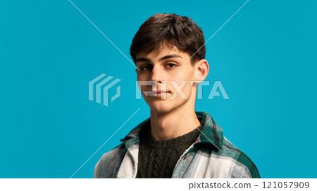 Close-up portrait of young handsome guy in checkered jacket, calmly posing, looking at camera against blue studio background 121057909