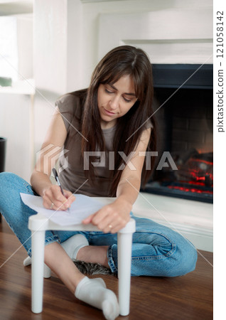 Young lady is seated at a kitchen table, attentively using a pen to write on a piece of paper writes a letter 121058142