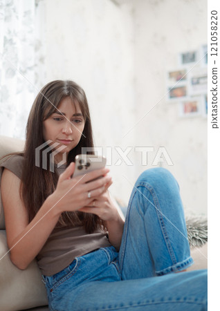Young happy woman sitting comfortably on sofa, smiling while using her phone indoors during a relaxing afternoon vertical 121058220