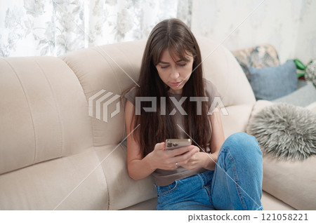 Young happy woman sitting comfortably on sofa, smiling while using her smart phone indoors during a relaxing afternoon Young happy woman sitting comfortably on sofa, smiling while using her smart phone indoors during a relaxing afternoon 121058221
