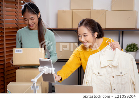 Two young asian woman live online clothing store while showing clothing with tablet and laptop. 121059204