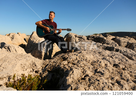 A man in a dark t-shirt playing acoustic guitar on a rocky area by the sea 121059400