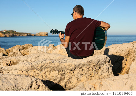 A man in a dark t-shirt playing acoustic guitar on a rocky area by the sea 121059405