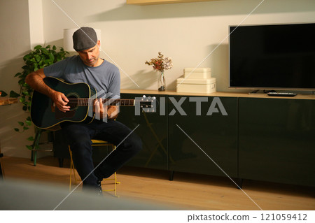 A man sitting at home learning to play a chord on acoustic guitar A man sitting at home learning to play a chord on acoustic guitar 121059412
