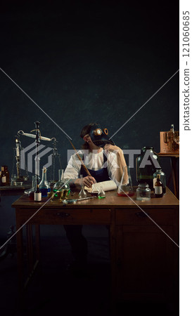Thoughtful man in plague doctor mask sits amidst vintage lab equipment, sits at table with feather in vintage laboratory against dark mystery background. 121060685