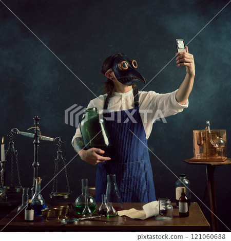 Holding large glass jar, the man in plague doctor mask takes modern selfie, hinting at his unconventional brewing or distilling work. Holding large glass jar, the man in plague doctor mask takes modern selfie, hinting at his unconventional brewing or distilling work. 121060688