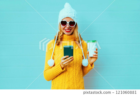 Winter portrait smiling young woman with phone and coffee cup in yellow sweater, white hat on blue 121060881