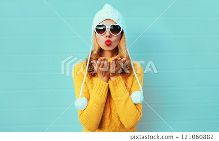 Winter portrait young woman blowing kiss wearing yellow knitted sweater and white hat on blue wall 121060882