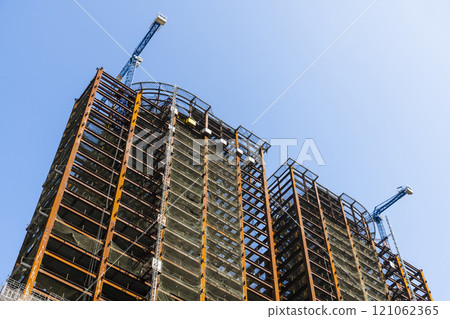 Low-angle view of cranes and steel structures of building construction with a blue sky background. 121062365