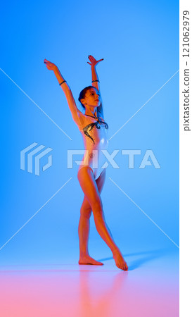 Artistic young girl, dressed festive shining suit, showing her athletic figure in pink neon light against gradient blue background. 121062979