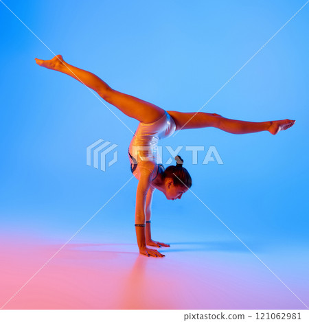 Young gymnast, flexible girl dressed festive costume performing handstand in pink neon light against gradient blue background. 121062981