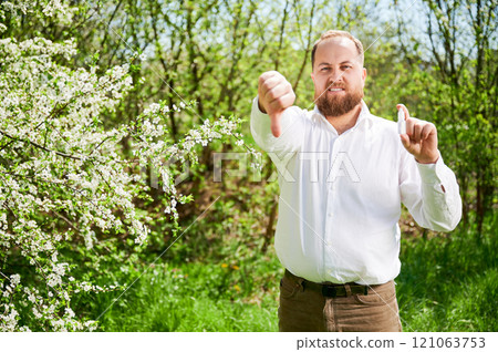 Man allergic using medical nasal spray, suffering from seasonal allergy at spring in blossoming garden. Handsome smiling man showing thumbs down near blooming tree outdoors. Spring allergy concept. Man allergic using medical nasal spray, suffering from seasonal allergy at spring in blossoming garden. Handsome smiling man showing thumbs down near blooming tree outdoors. Spring allergy concept. 121063753