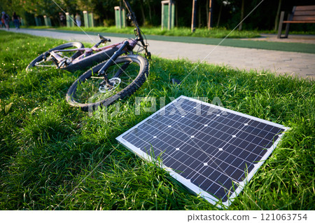 Pink electric mountain bike lies on lush green grass, connected to solar panel for charging. Bicycle beside paved path, showcasing bike's rugged tires and sleek design in outdoor environment. 121063754