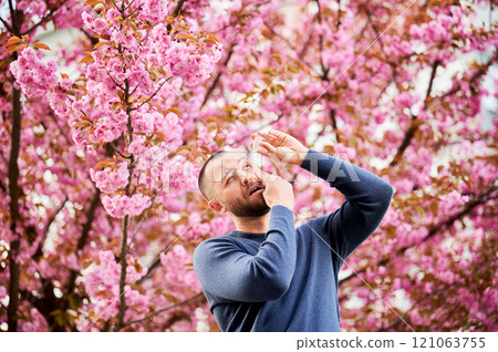 Man allergic using medical eyes drops, suffering from seasonal allergy at spring in blossoming garden. Handsome man treating eyes in front of blooming tree outdoors. Spring allergy concept. 121063755