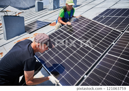 Workers building solar panel system on metal rooftop of house. Two men installers installing photovoltaic solar module outdoors. Alternative, green and renewable energy generation concept. 121063759