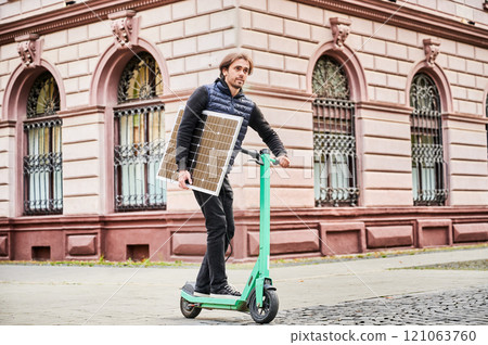 Man rides electric scooter while carrying solar panel. Integration of solar power as sustainable energy source for charging electric scooters, promoting eco-friendly urban transportation. Man rides electric scooter while carrying solar panel. Integration of solar power as sustainable energy source for charging electric scooters, promoting eco-friendly urban transportation. 121063760