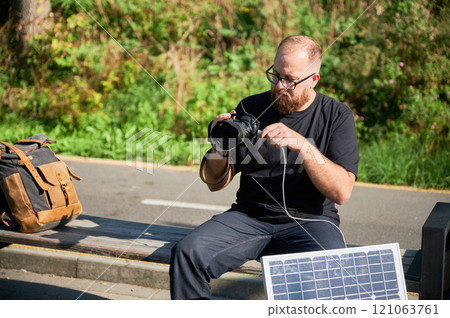 Bearded male photographer sits on bench, connecting camera to solar panel for charging. Integration of renewable energy into photography, using solar power to charge equipment during outdoor shoots. Bearded male photographer sits on bench, connecting camera to solar panel for charging. Integration of renewable energy into photography, using solar power to charge equipment during outdoor shoots. 121063761