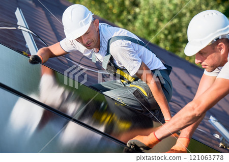 Workers building solar panel system on rooftop of house. Two men installers in helmets installing photovoltaic solar module outdoors. Alternative, green and renewable energy generation concept. 121063778