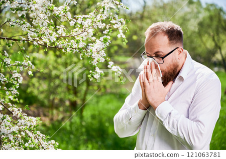 Man allergic suffering from seasonal allergy at spring in blossoming garden at springtime. Young man sneezing and blowing nose with nasal handkerchief in front of blooming tree. Spring allergy concept Man allergic suffering from seasonal allergy at spring in blossoming garden at springtime. Young man sneezing and blowing nose with nasal handkerchief in front of blooming tree. Spring allergy concept 121063781