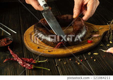 A skilled chef prepares to slice a flavorful sausage on a wooden cutting board. Fresh herbs and spices surround the dish, enhancing the culinary experience in a cozy kitchen setting 121064211
