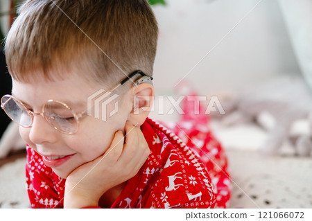 funny boy with hearing aids, glasses and red pajamas smiles widely while lying on a knitted rug on the floor funny boy with hearing aids, glasses and red pajamas smiles widely while lying on a knitted rug on the floor 121066072