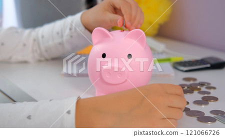 Child girl attentively placing coins into her piggy bank while sitting at her table at home. Personal finances and managing money in young age Child girl attentively placing coins into her piggy bank while sitting at her table at home. Personal finances and managing money in young age 121066726
