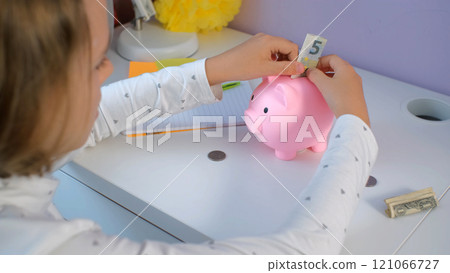 Young girl adding coins and banknotes into a pink piggy bank while sitting at desk. Personal finances and managing money Young girl adding coins and banknotes into a pink piggy bank while sitting at desk. Personal finances and managing money 121066727