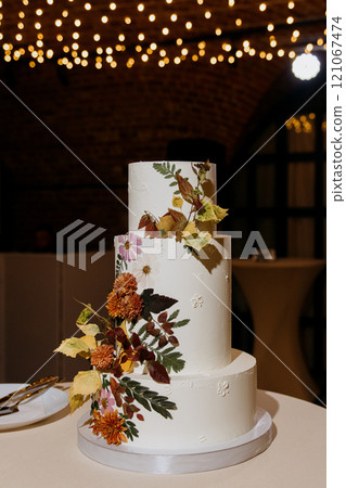 A wedding cake adorned with flowers and leaves sits on a table 121067474