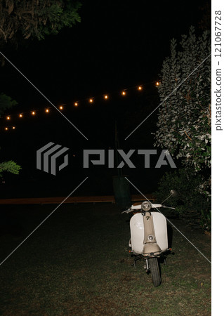 A white scooter is parked on the grass during the nighttime hours A white scooter is parked on the grass during the nighttime hours 121067728