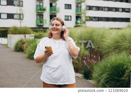 Happy young caucasian woman in good mood listening to music with headphones and standing on the street in the city. Music lover enjoying music. Urban lifestyle concept. Happy young caucasian woman in good mood listening to music with headphones and standing on the street in the city. Music lover enjoying music. Urban lifestyle concept. 121069020