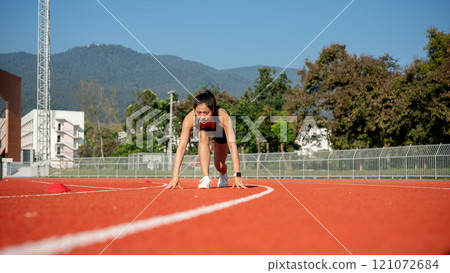 A determined woman sprinter in the starting position, ready to race on a racetrack at a stadium. 121072684