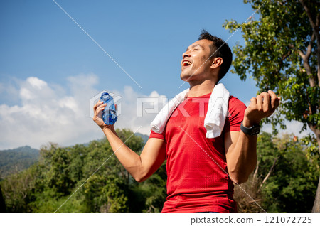 Happy Asian man in sportswear feels refreshed after drinking water following his workout in a park. 121072725