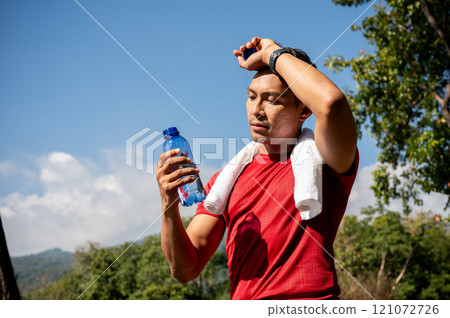 A sweaty, tired Asian man in sportswear drinks water while running in a park on a hot, sunny day. A sweaty, tired Asian man in sportswear drinks water while running in a park on a hot, sunny day. 121072726