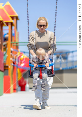 Mother pushing her infant baby boy child on a swing on playground outdoors Mother pushing her infant baby boy child on a swing on playground outdoors 121077721