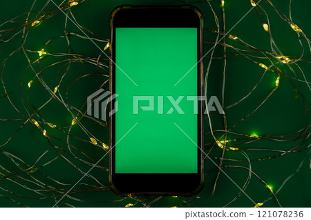 The phone lies horizontally with a green chromakey screen against a garland of yellow and green lights on a green background. High quality photo The phone lies horizontally with a green chromakey screen against a garland of yellow and green lights on a green background. High quality photo 121078236