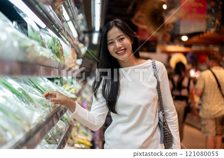 A smiling Asian woman grocery shopping at a supermarket, selecting fresh vegetables from a shelf. A smiling Asian woman grocery shopping at a supermarket, selecting fresh vegetables from a shelf. 121080055