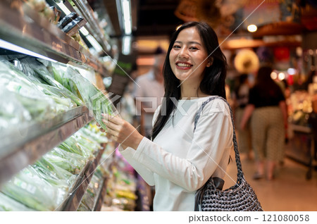 A smiling Asian woman grocery shopping at a supermarket, selecting fresh vegetables from a shelf. A smiling Asian woman grocery shopping at a supermarket, selecting fresh vegetables from a shelf. 121080058