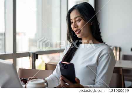 A woman holding her smartphone while working on her laptop computer, working in a modern office 121080069