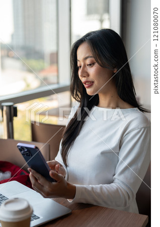 Attractive Asian woman reading messages on her smartphone while working remotely from a coffee shop. 121080070