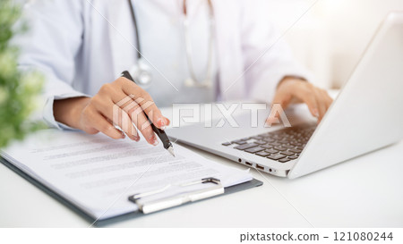 A close-up of a doctor working at her desk, typing on the laptop keyboard, reading medical paper. A close-up of a doctor working at her desk, typing on the laptop keyboard, reading medical paper. 121080244