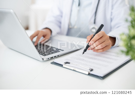 A close-up of a doctor working at her desk, typing on the laptop keyboard, reading medical paper. A close-up of a doctor working at her desk, typing on the laptop keyboard, reading medical paper. 121080245