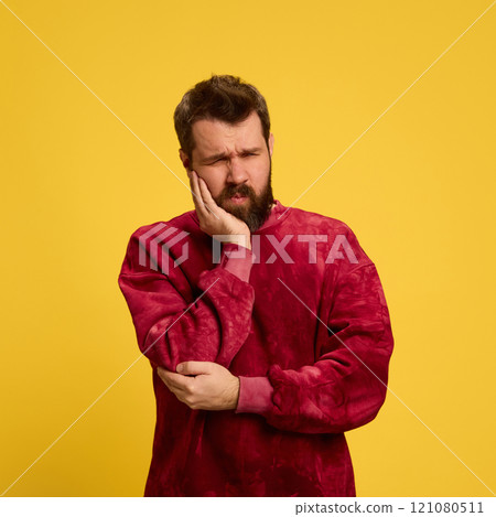 Young sad man, dressed red shirt holding hand on cheek as he feels tooth pain against bright yellow background. Viruses and symptoms. 121080511